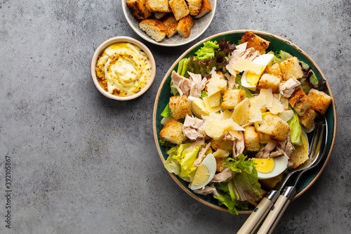 Fresh Caesar salad with lettuce salad, chicken breast, boiled eggs and croutons in ceramic bowl with dressing on the side on gray table. Classic healthy salad, top view, space for text