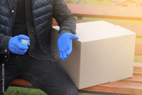 Delivery service courier during the Coronavirus, COVID-19, pandemic, cropped courier in medical protective mask, gloves spraying alcohol disinfectant spray on hands near cardboard boxes outdoors
