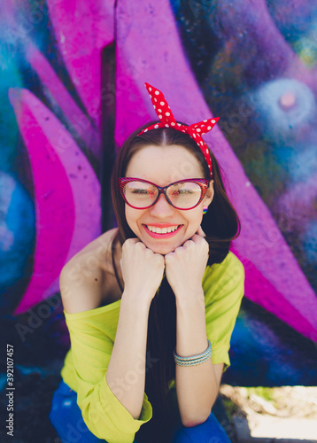 A stylish teenage girl in glasses and blue jeans poses near a graffiti wall