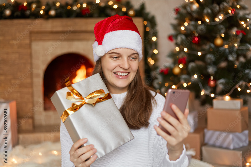 Photo of charming cute beautiful attractive girl holding her phone and gift box in hands, smiling female has video call or selfie with her Christmas present, posing in decorated living room. Photo of charming cute beautiful attractive girl holding her phone and gift box in hands, smiling female has video call or selfie with her Christmas present, posing in decorated living room.