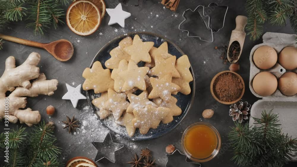 Top-down view, powdered sugar falling on traditional Christmas cookies. Gingerbread cookies in a plate and ingredients for the dough rotate on their axis. Christmas sweets background. Top-down view, powdered sugar falling on traditional Christmas cookies. Gingerbread cookies in a plate and ingredients for the dough rotate on their axis. Christmas sweets background.