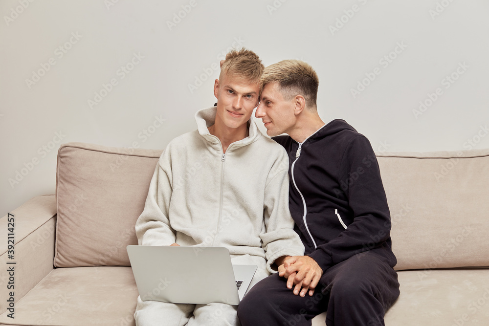 Cute gay couple sitting at home on the couch in their pajamas with a laptop  on their lap. LGBT couple. Stock Photo | Adobe Stock