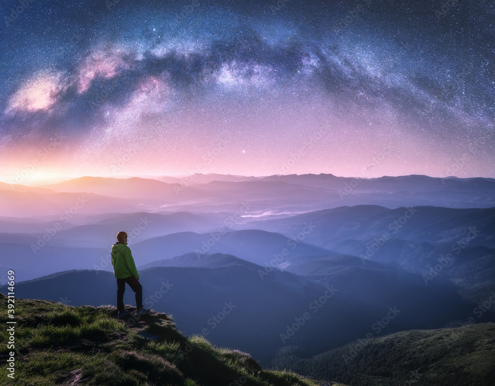 Man on the mountain peak and arched Milky Way over mountains in low clouds at night. Landscape with purple starry sky, Milky Way Arch, orange light, guy, hills in fog. Space and galaxy. Sky with stars