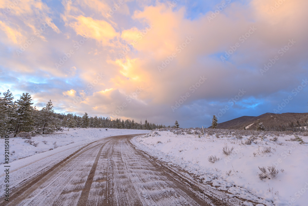 Vehicle tracks in snow on a dirt road near mountains and trees during winter with colorful clouds at sunset Vehicle tracks in snow on a dirt road near mountains and trees during winter with colorful clouds at sunset