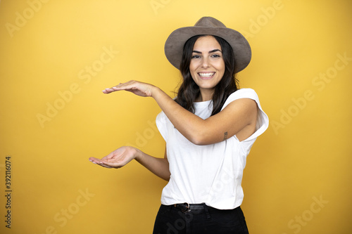 Beautiful woman wearing casual white t-shirt and a hat standing over yellow background gesturing with hands showing big and large size sign, measure symbol. Smiling . Measuring concept. © Irene