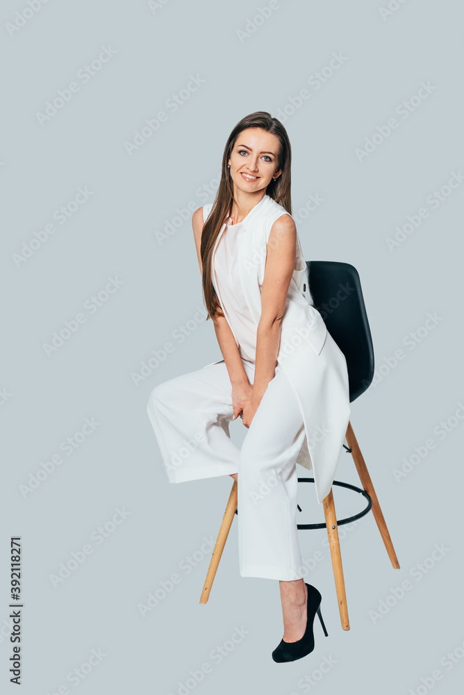 Pretty woman posing sitting in white suit on a chair over gray background. Woman with long dark hair posing to the camera. Pretty woman posing sitting in white suit on a chair over gray background. Woman with long dark hair posing to the camera.