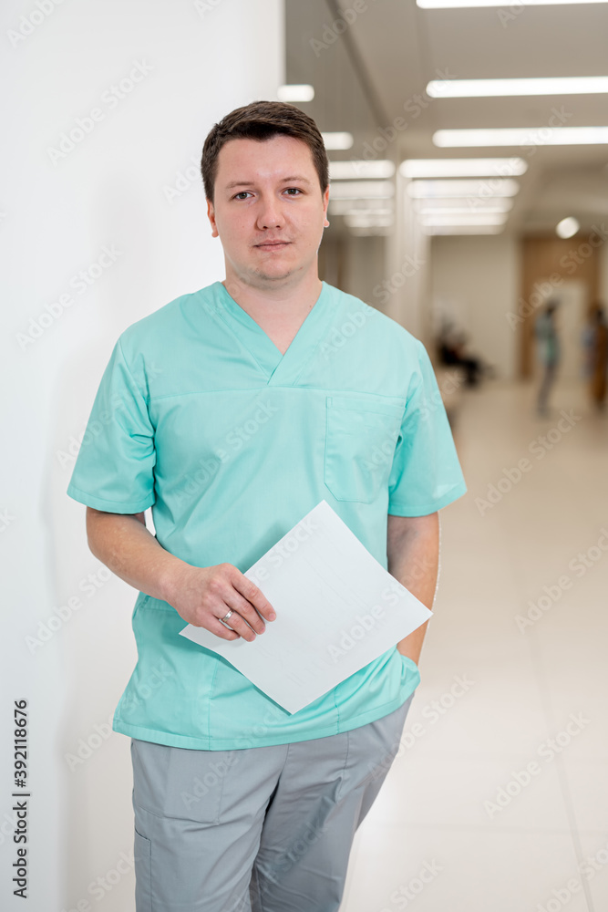 A male medical doctor in a uniform holding a clipboard and posing in a hospital corridor