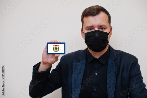 Man wear black formal and protect face mask, hold West Virginia flag card isolated on white background. USA coronavirus Covid country concept.