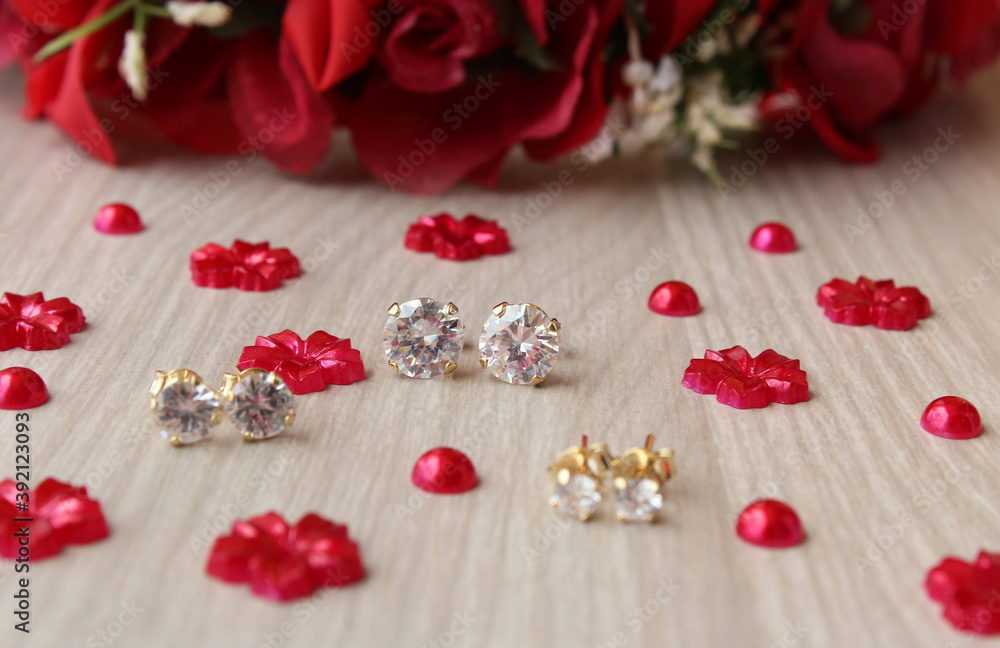 Brincos de ouro com pontos de luz de diamante/zircônia brilhantes sobre fundo de madeira com flores artificiais vermelhas.