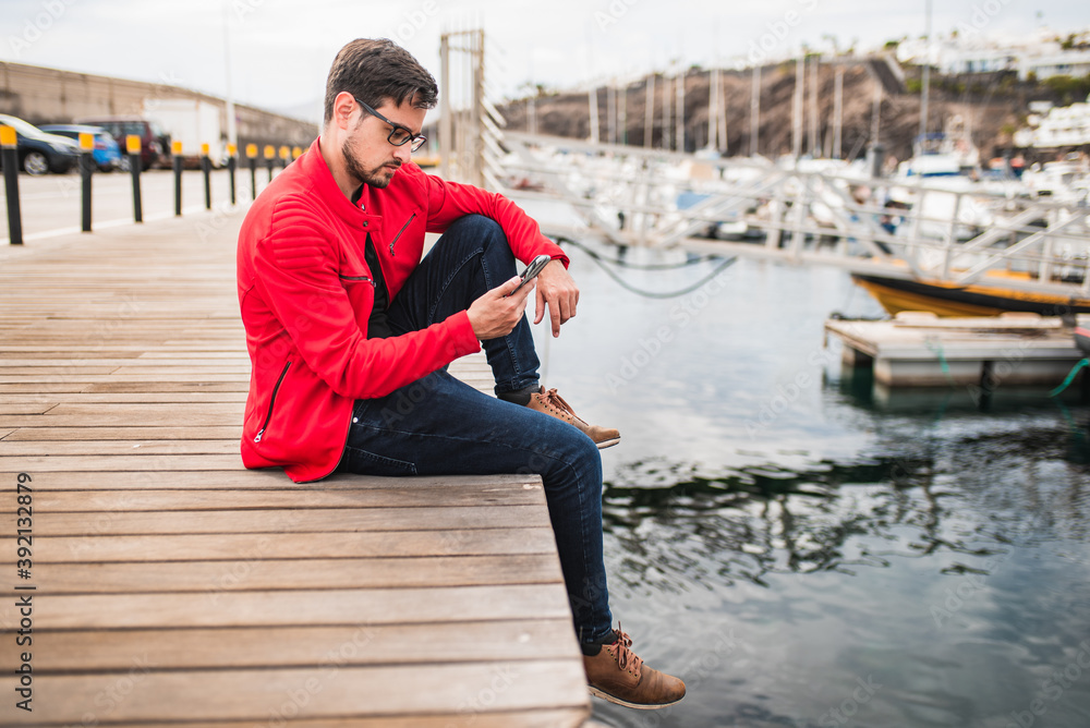 Young man sitting by the port consulting his smartphone