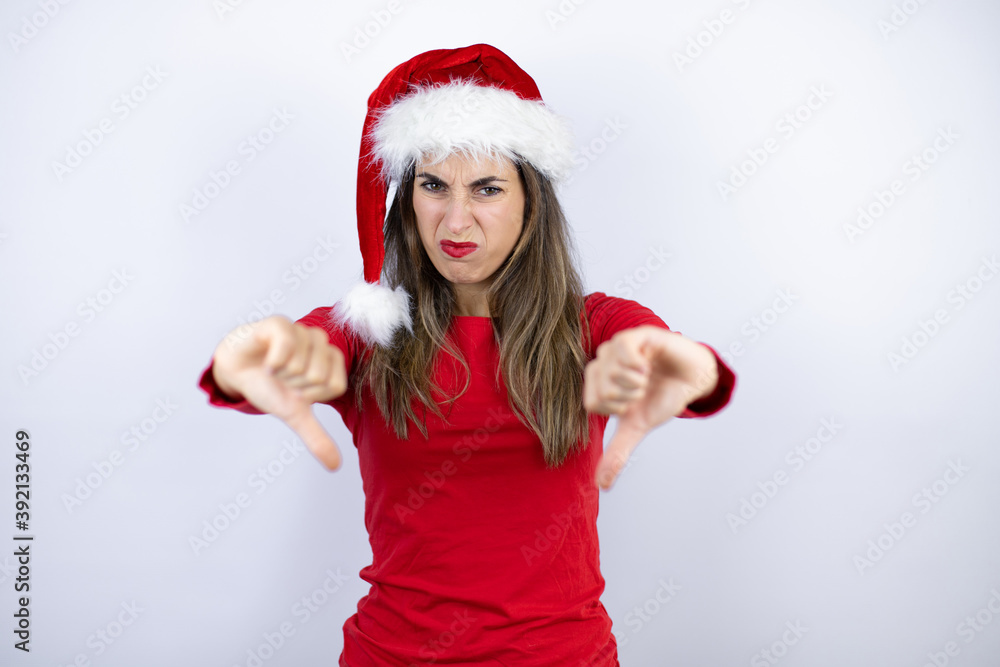 Young beautiful woman wearing a Santa hat over white background with angry face, negative sign showing dislike with thumbs down Young beautiful woman wearing a Santa hat over white background with angry face, negative sign showing dislike with thumbs down
