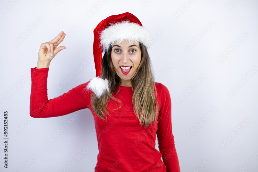 Young beautiful woman wearing a Santa hat over white background smiling and confident gesturing with hand doing small size sign with fingers . Measure concept. Young beautiful woman wearing a Santa hat over white background smiling and confident gesturing with hand doing small size sign with fingers . Measure concept.