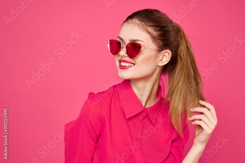 Cheerful woman in sunglasses touching hair with hands and pink shirt cropped view