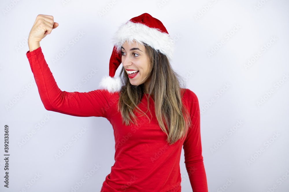 Young beautiful woman wearing a Santa hat over white background showing arms muscles smiling proud Young beautiful woman wearing a Santa hat over white background showing arms muscles smiling proud