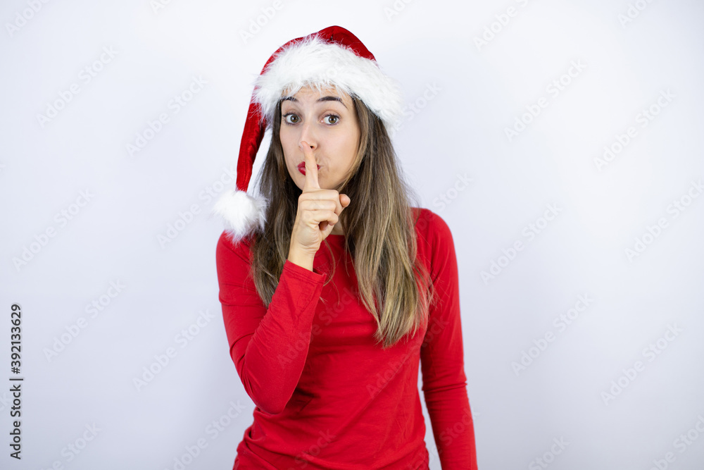 Young beautiful woman wearing a Santa hat over white background asking to be quiet with finger on lips. Silence and secret concept. Young beautiful woman wearing a Santa hat over white background asking to be quiet with finger on lips. Silence and secret concept.