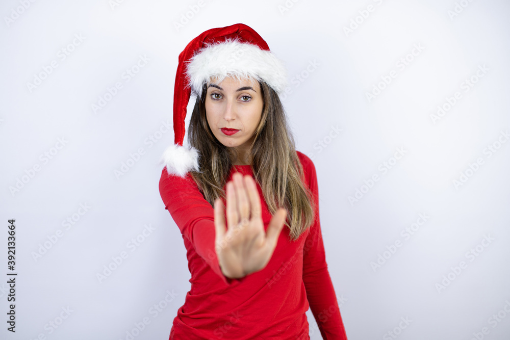 Young beautiful woman wearing a Santa hat over white background serious and doing stop sing with palm of the hand. Young beautiful woman wearing a Santa hat over white background serious and doing stop sing with palm of the hand.