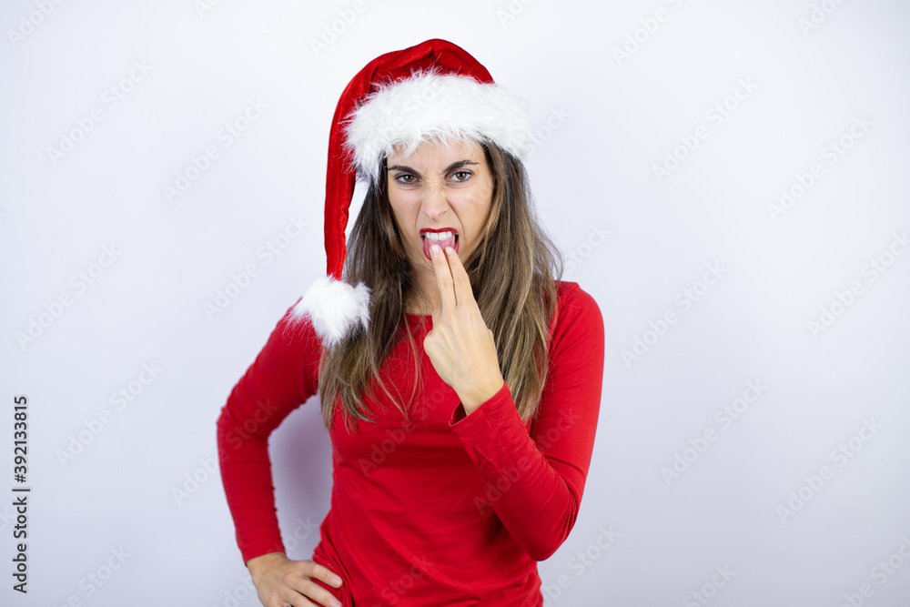 Young beautiful woman wearing a Santa hat over white background disgusted with her hand inside her mouth Young beautiful woman wearing a Santa hat over white background disgusted with her hand inside her mouth
