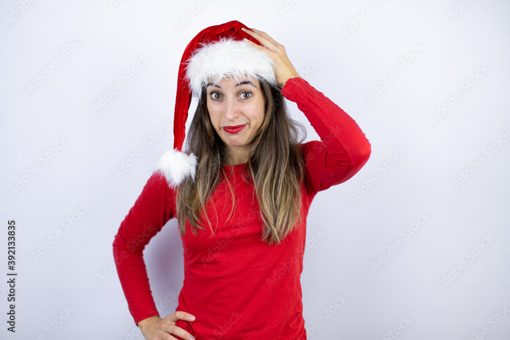 Young beautiful woman wearing a Santa hat over white background putting one hand on her head smiling like she had forgotten something Young beautiful woman wearing a Santa hat over white background putting one hand on her head smiling like she had forgotten something