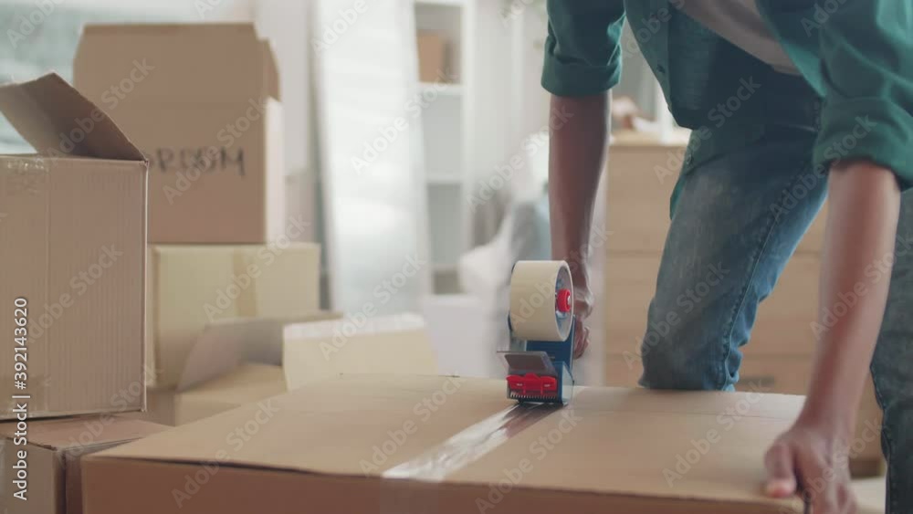 Lockdown of young African-American man holding packing machine and sticking duct tape on cardboard moving box Lockdown of young African-American man holding packing machine and sticking duct tape on cardboard moving box
