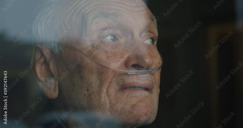 Cinematic close up shot of an elderly thoughtful man wearing oxygen tube is looking through a window in the morning. Concept of healthcare, life, pensioner, grandparents, retirement, boarding house.