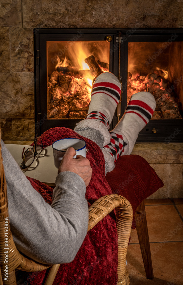 Winter night, happy woman resting by the fire with a blanket, a book, reading glasses and a cup of coffee, feet in wool socks. Cozy scene.