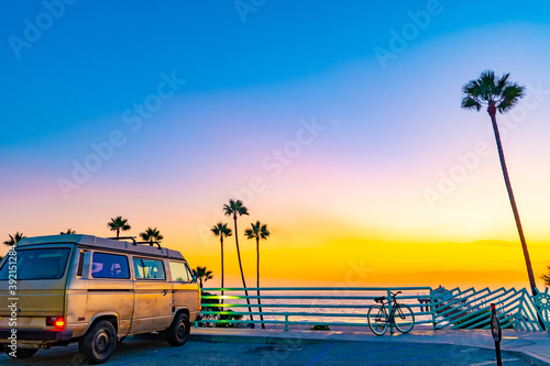 Scenic beach coast sunset view with tall California palm trees near the ocean and vintage van parked behind rail in street parking lot. Stunning sky colors at dusk. photo