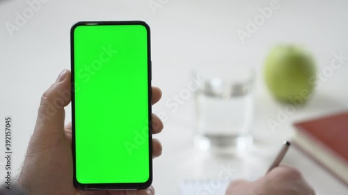 Close-up shot of green screen template smartphone in male hands at home offise, man is watcing content, makes notes in a notebook. Modent technology and information concept. photo