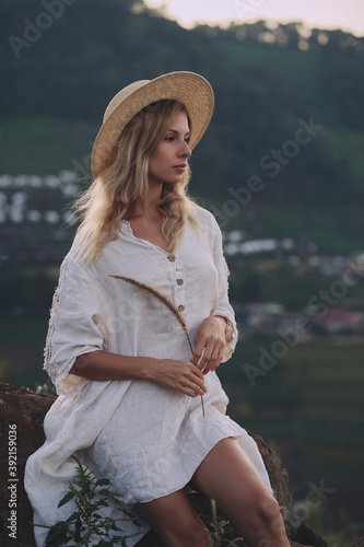 Obraz na płótnie The blonde in a linen dress and straw hat sits on a viewpoint in the mountains. A woman in the Boho style, resting in the countryside, a simple village lifestyle w salonie