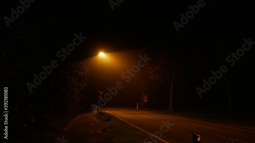 Einsame Landstra  e in der Nacht im Nebel mit einer fahlen Stra  enlaterne  Natriumdampf  erzeugt das typische Gef  hl der Einsamkeit in der Corona Covid19 Pandemie