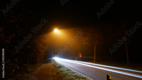 Einsame Landstraße in der Nacht im Nebel mit einer fahlen Straßenlaterne (Natriumdampf) und Leuchtspuren eines Autos durch Langzeitbelichtung