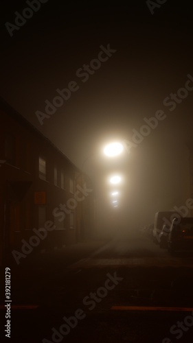 Blick in eine enge Gasse in der Nacht im dichten Nebel mit Strassenlaternen, die trübe Lichtinseln erzeugen und eine unheimliche, düstere Atmosphäre