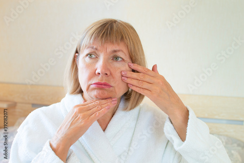 Portrait of a sad elderly woman background of his bedroom. Her hands touch the wrinkles on her face. Concept Anti aging, healthcare and cosmetology, mature people