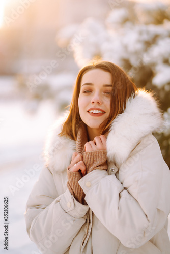 Portrait of young beautiful woman in a snowy park. Cheerful lady in winter clothes posing with joy outside in the snow forest. Winter holiday, Christmas, New year.