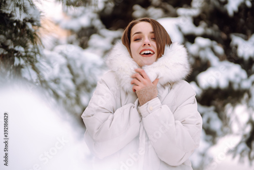 Portrait of young beautiful woman in a snowy park. Cheerful lady in winter clothes posing with joy outside in the snow forest. Winter holiday, Christmas, New year.