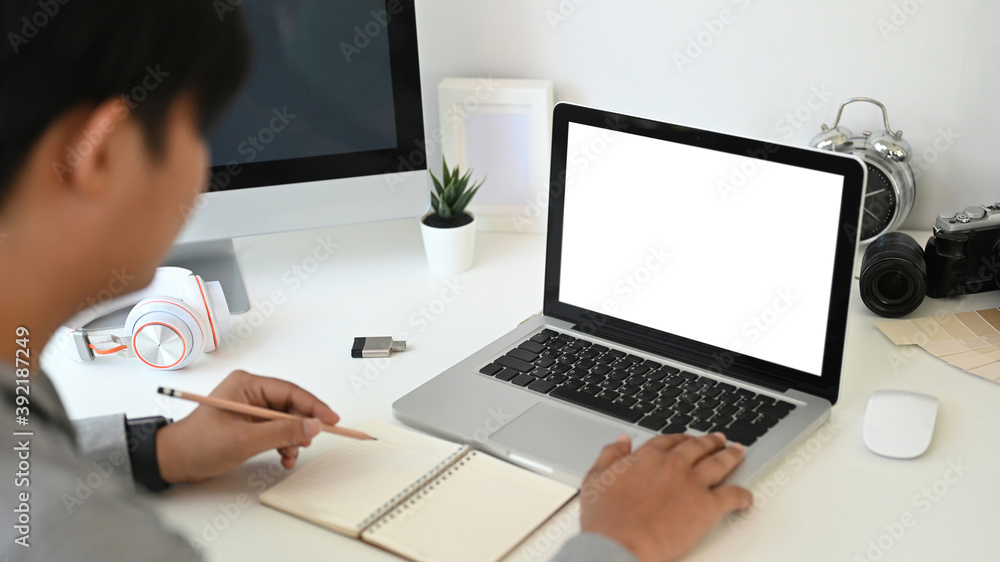 Cropped shot of young man freelancer is sitting in front of empty screen laptop and writing information on notebook. Cropped shot of young man freelancer is sitting in front of empty screen laptop and writing information on notebook.
