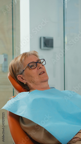 Close up of patient wearing dental bib talking with dentist before intervetion. Orthodontist speaking to woman with toothache sitting on stomatological chair while nurse preparing sterilized tools. photo