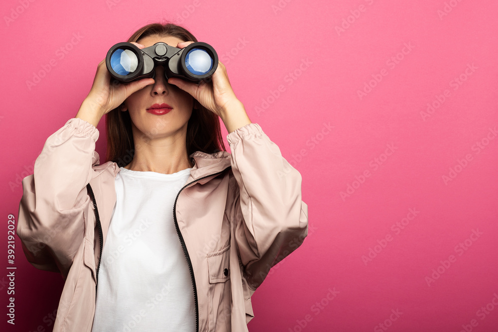 Young woman in jacket looking through binoculars on pink background. Young woman in jacket looking through binoculars on pink background.