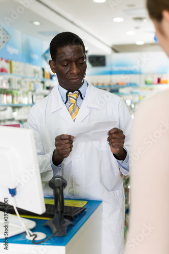 Professional African American male pharmaceutist reading prescription of client in modern pharmacy