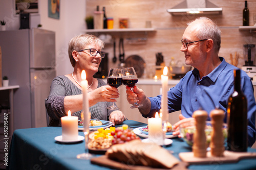 Elderly couple holding wine glasses while dining together in kitchen. Happy cheerful senior couple dining together in the cozy kitchen, enjoying the meal, celebrating their anniversary.
