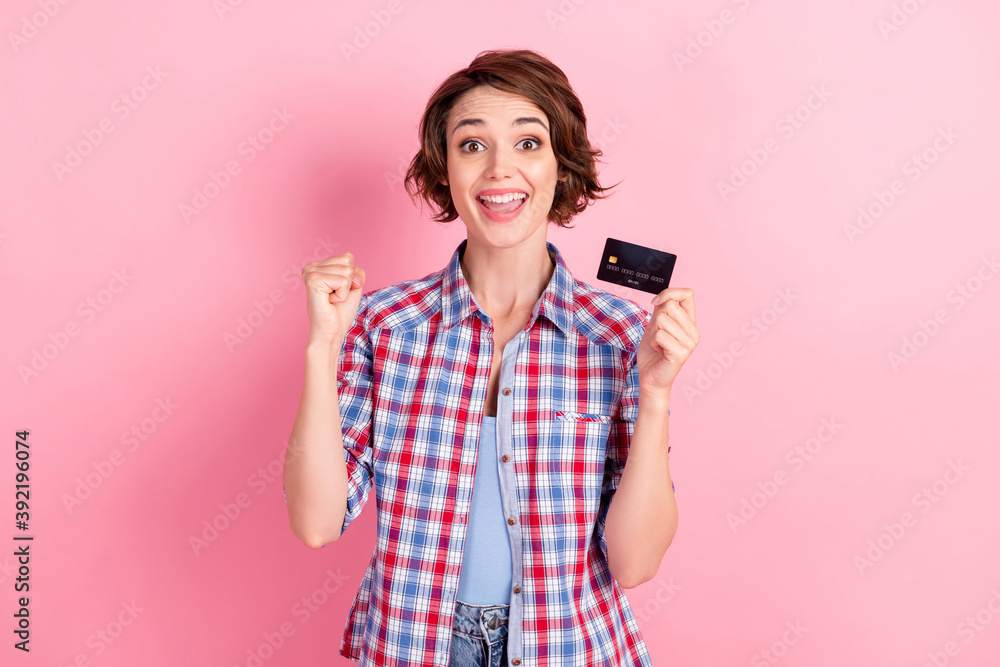 Photo of beaming lucky woman wear checkered shirt holding bank card hand arm fist isolated pink color background Photo of beaming lucky woman wear checkered shirt holding bank card hand arm fist isolated pink color background