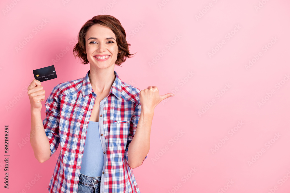 Photo of shiny toothy woman wear checkered shirt holding hand arm bank card thumb finger pointing empty space isolated pink color background Photo of shiny toothy woman wear checkered shirt holding hand arm bank card thumb finger pointing empty space isolated pink color background