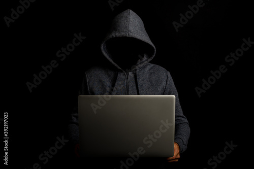 Man without a face in a hood holds a laptop on a dark background. Concept of cyber rogue, hacker. Banner