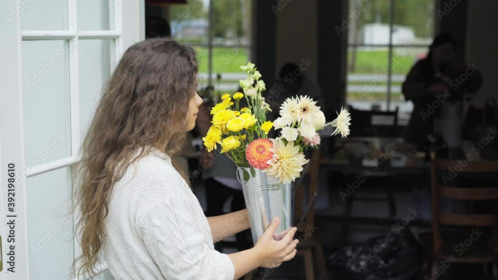 Pretty long haired woman in white blouse smells fresh colorful flowers in high metal bucket vase coming into dark room from house yard on spring day Pretty long haired woman in white blouse smells fresh colorful flowers in high metal bucket vase coming into dark room from house yard on spring day