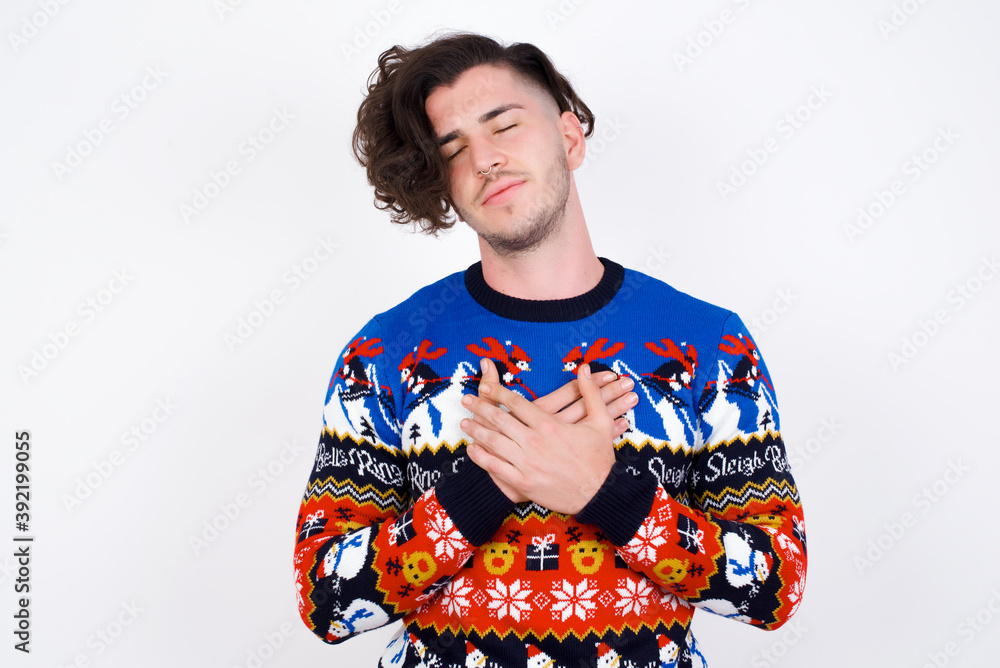 Young handsome Caucasian man wearing Christmas sweater closes eyes and keeps hands on chest, expresses sincere emotions, being kind hearted and honest. Body language and real feelings concept. Young handsome Caucasian man wearing Christmas sweater closes eyes and keeps hands on chest, expresses sincere emotions, being kind hearted and honest. Body language and real feelings concept.