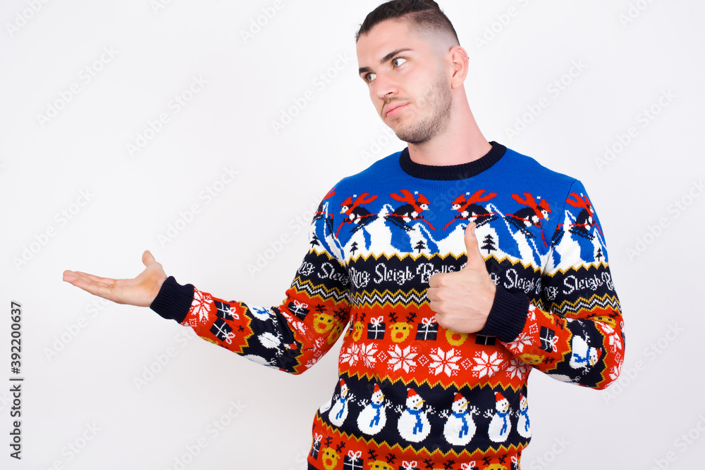 Happy cheerful Young handsome Caucasian man wearing Christmas sweater against white wall showing thumb up and pointing with the other hand. I recommend this. Happy cheerful Young handsome Caucasian man wearing Christmas sweater against white wall showing thumb up and pointing with the other hand. I recommend this.