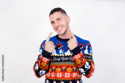 Strong healthy straight white teeth. Close up portrait of happy Young handsome Caucasian man wearing Christmas sweater against white wall with beaming smile pointing on perfect clear white teeth.