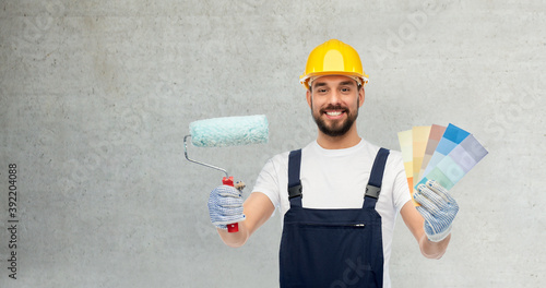profession, construction and building concept - happy smiling male worker or builder in yellow helmet and overall with paint roller and color palettes over grey concrete background photo