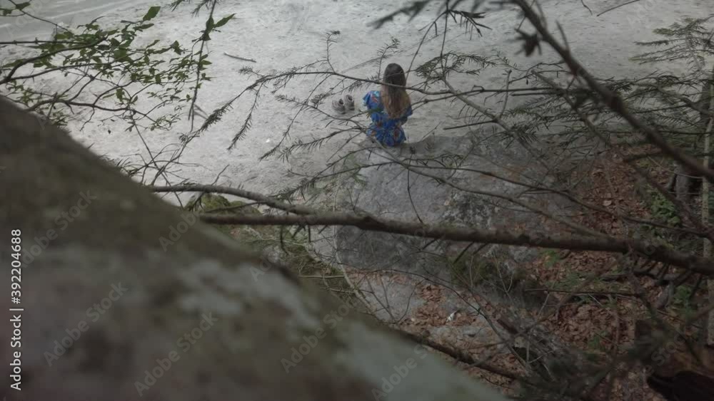 Romantic girl sitting on a rock in the forest with trees. Cool footage from above in the nature. Young adult woman with long air on a rock in a park. Aerial view from the back. Trentino, Italy. Romantic girl sitting on a rock in the forest with trees. Cool footage from above in the nature. Young adult woman with long air on a rock in a park. Aerial view from the back. Trentino, Italy.