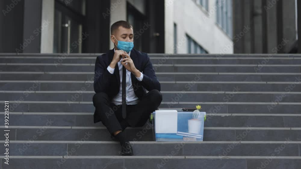 Fired male office worker in medical mask sitting on stairs in depression with box of stuff. Anxious concept. Workless man in despair. Unemployed businessman lost his business Fired male office worker in medical mask sitting on stairs in depression with box of stuff. Anxious concept. Workless man in despair. Unemployed businessman lost his business
