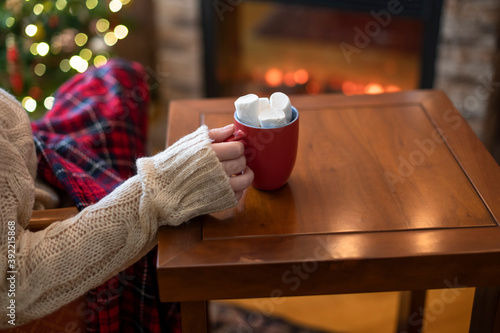 Woman with cup of hot cocoa and marshmallow sitting and warming at winter evening near fireplace flame, covered christmas plaid